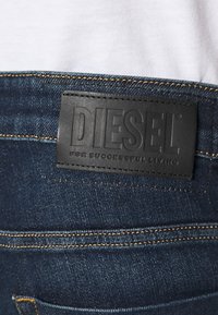 Mörkblå denimjeans med en svart läderlapp där "DIESEL" är präglat, samt sömmar i en kontrasterande guldfärg.