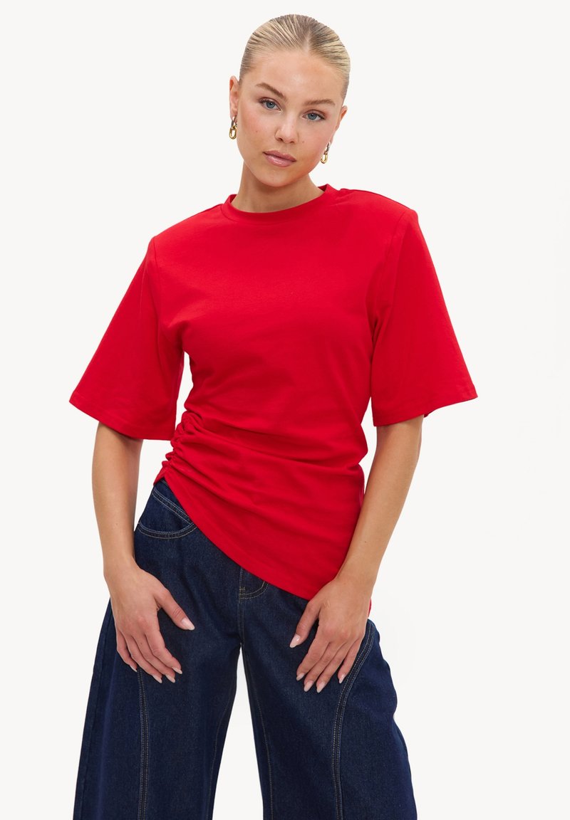 Femme aux cheveux blonds tirés en arrière, portant des boucles d'oreilles créoles dorées, un haut rouge à manches courtes froncé, et un jean large bleu foncé, debout sur un fond blanc.