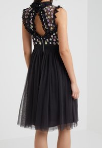 Vestido negro con un cuerpo ajustado bordado de flores y espalda transparente, cuello con volantes y una falda de tul fluida. Cremallera en la parte posterior.