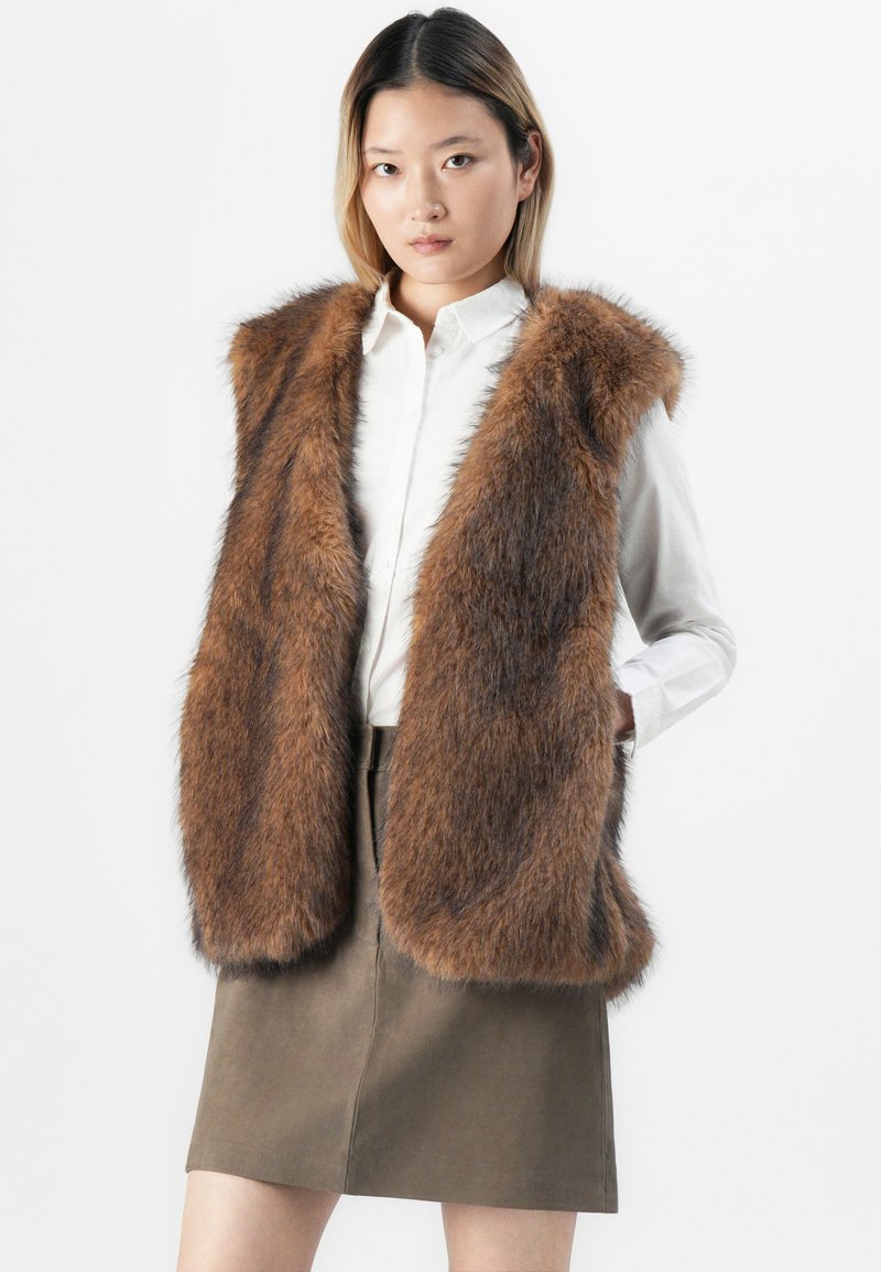 Donna che indossa un gilet in pelliccia sintetica marrone sopra una camicia bianca con bottoni e una gonna verde oliva, in piedi contro uno sfondo bianco.