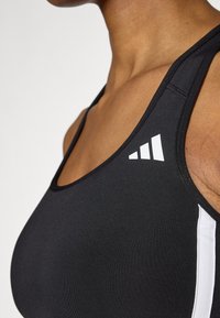 Μαύρο αθλητικό σουτιέν με λευκό λογότυπο Adidas, με κομψό σχέδιο, λεία υφή, λεπτές ράντες και αντίθετες λευκές πλευρικές γραμμές.