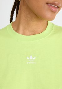 Svetlozelené bavlnené tričko s okrúhlym výstrihom a malou bielou vyšívanou značkou Adidas na hrudi. Hladká textúra.