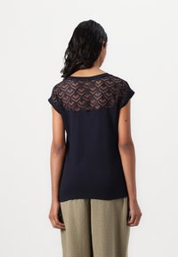 Marinblå t-shirt med korta ärmar och en genomskinlig spetsbakgrund med geometriskt mönster. Mjuk tyg, avslappnad passform, kombineras med strukturerade byxor.