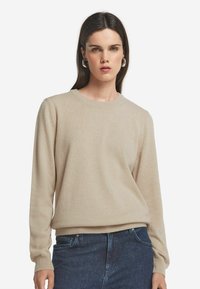 Beżowy sweter z miękkiej dzianiny, z okrągłym dekoltem, długimi rękawami i ściągaczami przy mankietach. W połączeniu z ciemnoniebieskimi dżinsami.