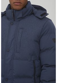 Navy blue puffer jacket with a textured finish, high collar, front button closure, zip pockets, and a logo patch on the shoulder.