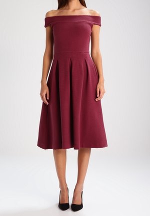 Žena v bordó šatech s odhalenými rameny, kolenní délky a plisovanými, která má na nohou černé lodičky, stojí na jednobarevném světlém pozadí.