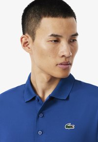 Blauw poloshirt van soepel materiaal, met een kraag en een knoopsluiting met drie knopen. Inclusief een klein geborduurd logo op de borst.