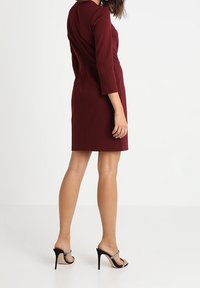 Burgundowa dopasowana sukienka z rękawami o długości trzech kwart, gładki materiał, do kolan, w połączeniu z czarnymi sandałami na wysokim obcasie z srebrnymi akcentami.