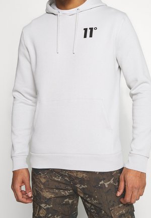 Homme portant un sweat à capuche blanc avec un logo noir "11°" sur la poitrine et un pantalon cargo camouflage marron, debout les bras détendus le long du corps.
