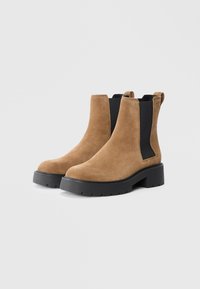 Brown suede ankle boots with black elastic side panels, chunky black sole, and logo detail on the heel. Smooth texture and rounded toe.