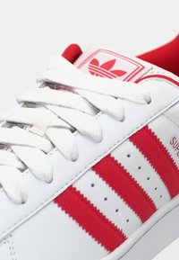 Fehér tornacipő piros három csíkkal és logóval. Simított bőr felsőrésszel, szövetből készült fűzőkkel és párnázott szegéllyel a kényelemért.