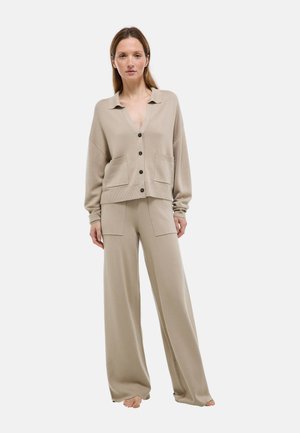 Femme portant un cardigan beige boutonné avec des poches et un pantalon large assorti, debout pieds nus devant un fond uni.