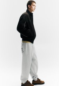 Jachetă bomber neagră cu margini albe, pantaloni de trening gri cu buzunar lateral și detaliu imprimat, și pantofi maro cu textură. Fundal simplu.