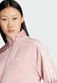 Ružová športová vetrovka na zips s vysokým golierom; obsahuje biele textilné pruhy pozdĺž ramien a malé biele logo na hrudi.