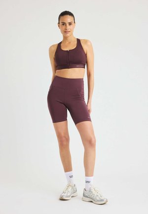 Donna in reggiseno sportivo bordeaux e pantaloni a ciclismo con calze bianche e sneaker su uno sfondo chiaro e semplice.