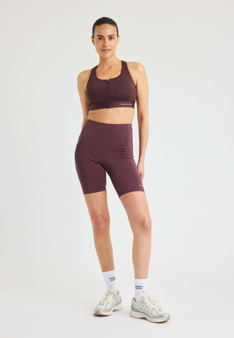 Donna in reggiseno sportivo bordeaux e pantaloni a ciclismo con calze bianche e sneaker su uno sfondo chiaro e semplice.