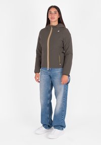 Jonge vrouw staat rechtop, gekleed in een bruine hoodie met rits, losse blauwe spijkerbroek en witte sneakers, tegen een eenvoudige witte achtergrond.