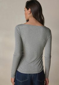 Graues Langarmoberteil aus weichem, leichtem Stoff mit einem Bootsausschnitt. Die Rückseite hat eine glatte Textur und wird mit dunkelblauer Jeans kombiniert.