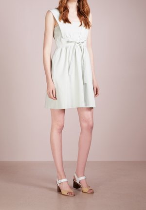 Woman wearing a light-colored sleeveless dress with a waist tie and beige and white heeled sandals, standing against a plain background.