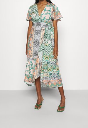 Femme portant une robe portefeuille patchwork verte, pêche et blanche à manches courtes, avec des sandales vertes à bouts ouverts, debout sur un sol gris.