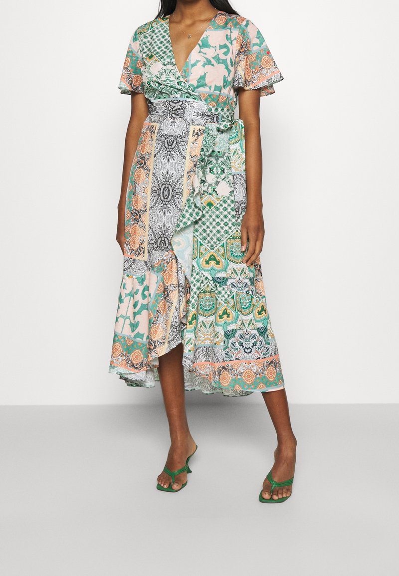 Femme portant une robe portefeuille patchwork verte, pêche et blanche à manches courtes, avec des sandales vertes à bouts ouverts, debout sur un sol gris.