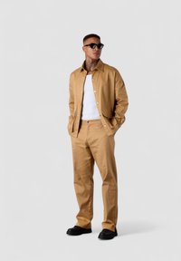 Tan jacket and matching pants made of smooth fabric, worn over a white tank top. Black sunglasses and shoes complete the look.