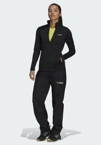 Giacca sportiva nera con zip frontale, maniche lunghe e logo Terrex. Abbinata a pantaloni neri e scarpe da trekking nere.