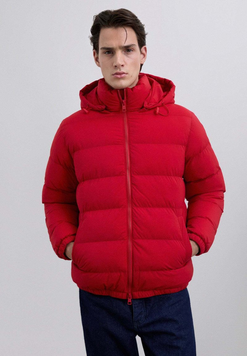 Joven con una chaqueta acolchada roja brillante con capucha, con las manos en los bolsillos, de pie frente a un fondo gris claro liso.