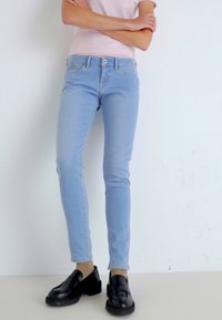 Světle modré džíny vyrobené z denimu, které mají úzký střih a jemné vyblednutí, v kombinaci s černými mokasíny. Textura vypadá hladce, s klasickým prošíváním.
