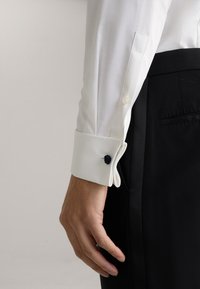 Weißes Hemd mit glattem Stoff, Navy-blauer Manschettenknopf, detaillierte Nähte, getragen mit schwarzen Hosen.