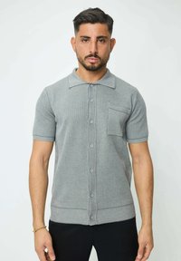 Polo en tricot gris à manches courtes avec une fermeture à boutons, un col et une poche poitrine. Présente des poignets et un ourlet côtelés pour un look ajusté.