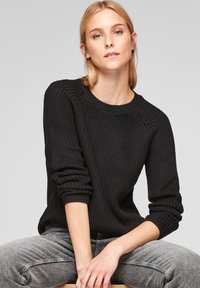Pull en maille noire avec un motif texturé, encolure ras du cou côtelée, et manches longues, associé à un jean gris. Design simple et décontracté.