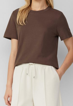 Kobieta w brązowym t-shircie z krótkim rękawem wpuszczonym w kremowe spodnie z wysokim stanem i wiązanym paskiem, jedną ręką w kieszeni.