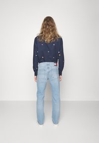 Camisa navy con bordado geométrico multicolor, combinada con jeans azul claro, que presenta una etiqueta en la parte trasera de la cintura y un diseño de pierna recta.