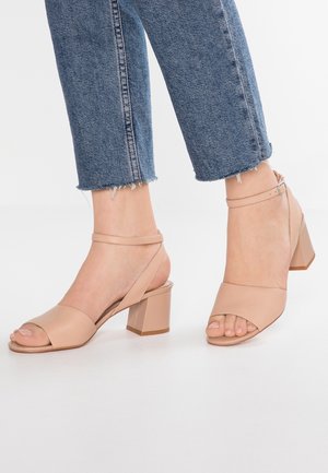 Sandalias de tacón abierto en color beis con tacón cuadrado, que cuentan con un diseño de tiras cruzadas y una correa en el tobillo, combinadas con pantalones de mezclilla cortos.