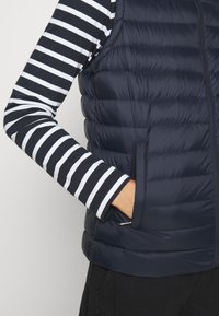 Granatowa kamizelka puchowa z poziomym pikowaniem, z zamykanymi kieszeniami i kontrastującymi paskami na rękawach w biało-granatowym kolorze.