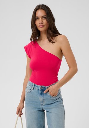 Vrouw draagt een felroze top met één schouder, lichtblauwe jeans met één hand in de zak, houdt een dun koordriempje vast in de andere hand.