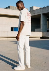 Moški v belih hlačah in supergah nosi bež majico z rdečim napisom "Slow Sunday Coffee Club" in grafiko skodelice na hrbtu, stoji na prostem.