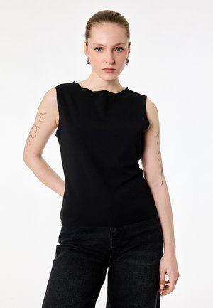 Giovane donna con capelli tirati indietro indossa un top nero senza maniche e pantaloni neri, mostrando tatuaggi su entrambe le braccia superiori su sfondo bianco.