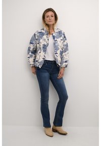 Vit bomberjacka med blå blommönster, ribbade manschetter, rund halsringning; kombinerad med blå jeans och beige ankelstövlar. Slät textur.