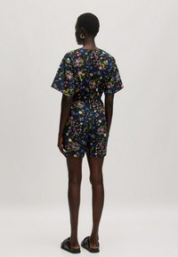 Blumenmuster-Short-Romper aus schwarzem Stoff, mit kurzen Ärmeln, elastischem Taillenbund und einer Mischung aus bunten Blumenmustern. Flache Sandalen getragen.
