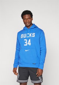 Mėlynas džemperis su užrašu "BUCKS" ir numeriu "34" balta spalva, Nike logotipas ant kišenės, derinamas su juodais sportiniais šortais, kurie turi baltas juostas.