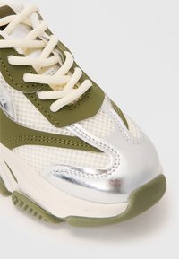 Basket en maille verte olive et blanche, avec des accents argentés, des lacets crème et une semelle en caoutchouc texturée.
