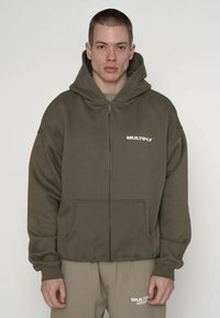 Sweat à capuche zippé vert olive avec une poche kangourou, des épaules tombantes, et un logo blanc imprimé « MULTIPLY » sur le côté gauche de la poitrine.