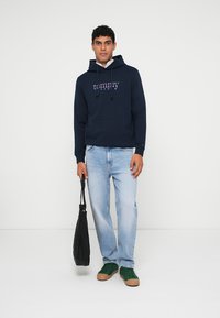 Navy hoodie with "tommy" logo, drawstrings, and front pocket. Light blue jeans and green sneakers. The model holds a black bag.