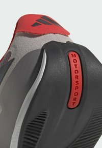 Close-up van de zool van een grijze en rode sneaker met een zwart profiel en rode tekst "MOTORSPORT" op de hiel.