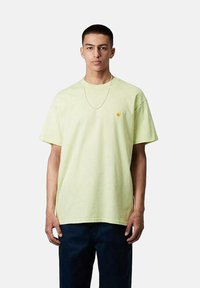 Camiseta de algodón amarillo claro con mangas cortas, corte relajado y un pequeño logo naranja en el pecho. Llevada con pantalones azul marino oscuro.