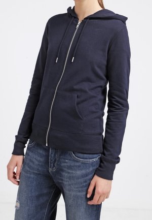 Personne portant un sweat à capuche bleu marine zippé avec une poche devant et des cordons, assorti à un jean bleu déchiré.