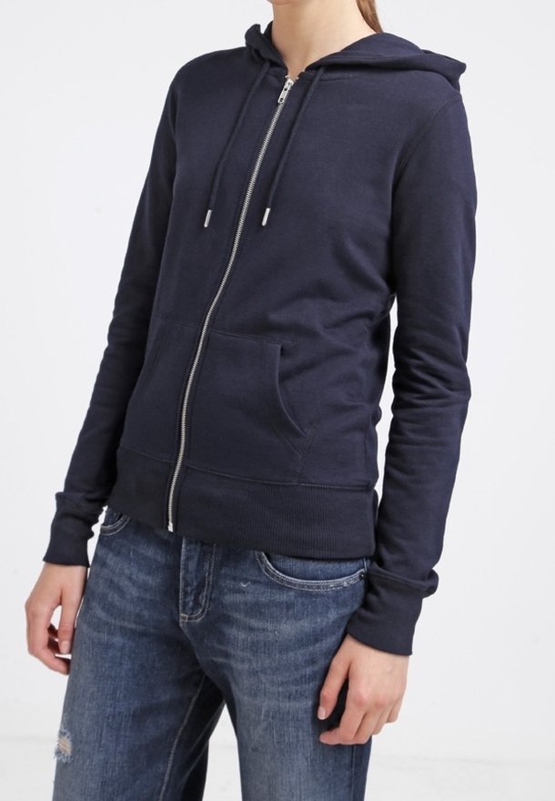 Personne portant un sweat à capuche bleu marine zippé avec une poche devant et des cordons, assorti à un jean bleu déchiré.
