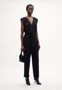 Mono negro con mangas cortas, escote en V profundo, cinturón en la cintura y puños enrollados. La modelo sostiene un pequeño bolso negro. Los tacones de aguja completan el look.